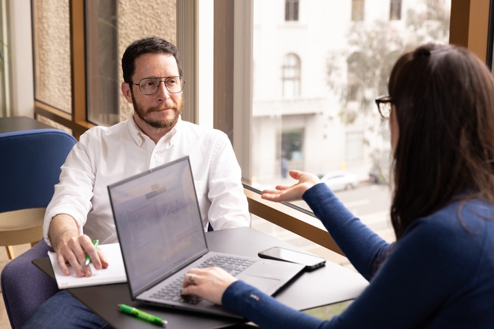 Two people in a modern office setting are having a conversation across a table with a laptop open. A woman, facing away from the camera, is gesturing while speaking to a man in glasses who appears to be listening attentively and taking notes on a notepad. A large window with a cityscape view is in the background.