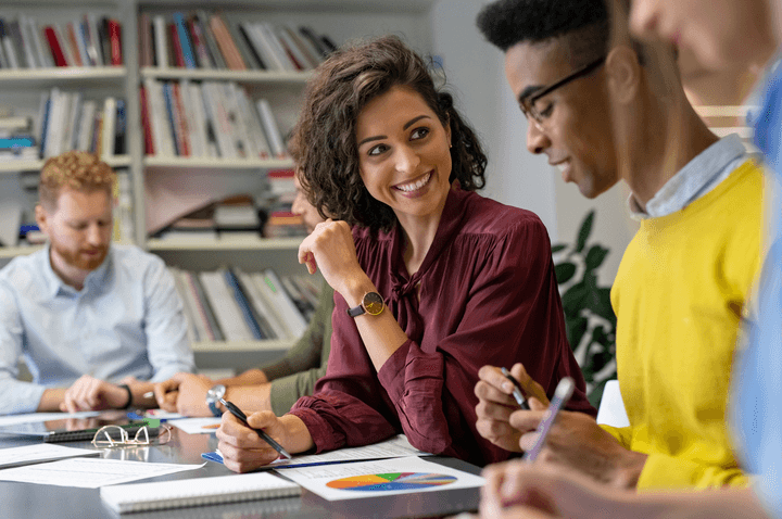 A diverse group of people engaged in a meeting at an office table. A woman in a burgundy blouse is smiling while interacting with her colleagues. They are discussing a pie chart document, with a bookshelf filled with books in the background.