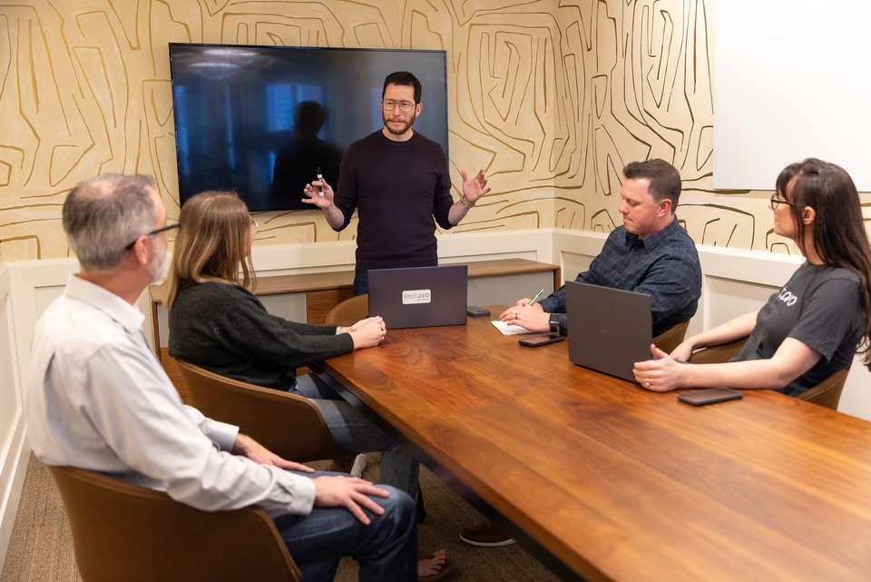 A man stands and gestures while speaking to four seated people at a conference table, with laptops and notebooks in front of them. A large screen is visible behind the speaker.