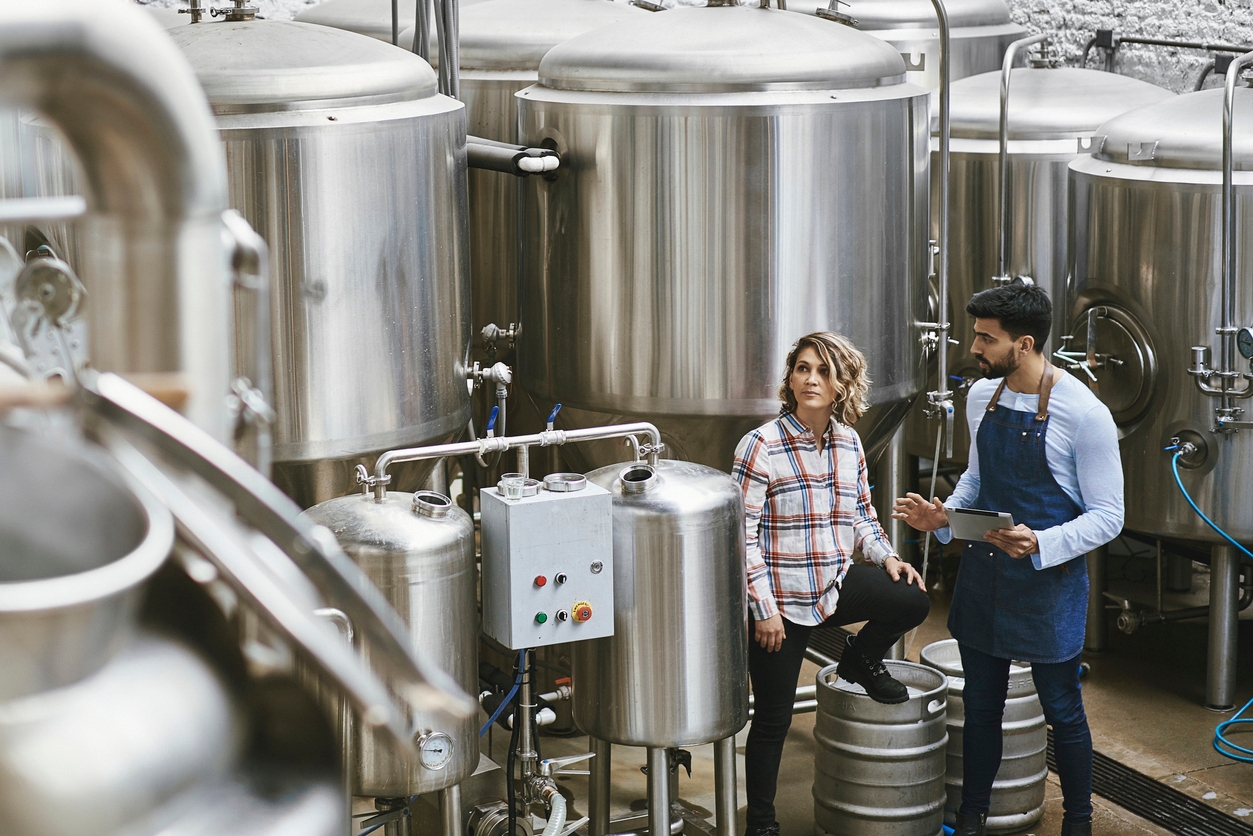 
                  A man and a woman conversing inside a brewery with large stainless steel fermentation tanks in the background. The man, in a denim apron and holding a tablet, appears to be discussing brewing processes with the woman, who is casually sitting on a metal keg.
                